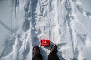 A person is standing in the fresh snow, with a first aid kit carefully placed on the ground beside them, ready for any possible injuries that may occur while enjoying the winter outdoors photo