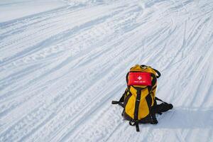 A bright yellow backpack sits prominently in the snow, accompanied by a striking red first aid kit, highlighting the adventure and preparations needed for winter activities photo