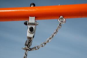 A detailed closeup view featuring a sturdy metal hook and robust chain mounted on a vibrant orange frame, set against a backdrop of a clear blue sky, showcasing industrial design photo