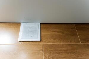 A white vent is strategically located on a beautifully arranged and polished wooden floor right next to a wall, creating a visually appealing contrast within the surrounding environment photo
