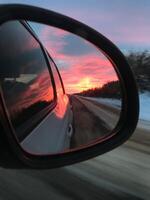 As the sun gradually sets on the horizon, its vibrant and warm colors beautifully reflect in a cars rearview mirror, creating a truly stunning visual experience for all drivers on the road photo