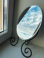 A beautifully crafted round mirror, made with exquisite materials, is placed on a window sill, reflecting the soft, warm natural light streaming in from the charming window photo