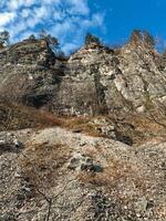 A beautifully rugged and rocky hillside adorned with trees and vegetation, all set against a bright blue sky in the background that enhances its picturesque scenery, inviting exploration photo