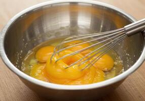 Several raw eggs are in a stainless steel bowl, with a whisk poised to mix them for baking. photo