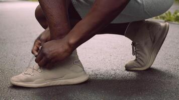 A man is crouched down, tying his shoelaces in a public park. He is dressed in athletic wear, ready to embark on an outdoor exercise routine, enjoying a healthy lifestyle. video