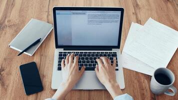 A person is focused on typing on a laptop, surrounded by papers and a coffee cup on a wooden desk, indicating a productive work session in a cozy environment. video