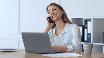 A woman smiles as she speaks on her phone in a bright office setting. She is seated at a desk with a laptop open, surrounded by office supplies and a coffee cup. video