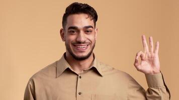 A cheerful Middle Eastern man stands in front of a neutral backdrop, confidently displaying the OK hand gesture. His expression conveys positivity and assurance, emanating a friendly lifestyle vibe. video