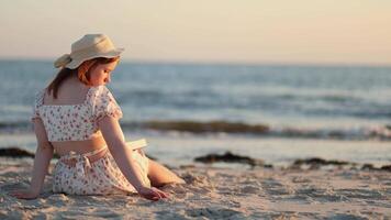A young woman enjoys a peaceful evening by the shore, reading a book while the sun sets on a serene beach video