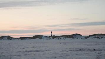 Sunset over the snowy dunes with a lighthouse standing tall in the distance on a serene winter evening video