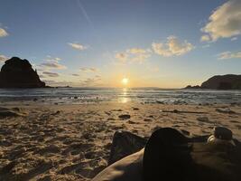 Sunset view of Papuma Beach in Jember, featuring a rocky islet standing tall against the warm hues of the setting sun. The sky transitions from deep blue to orange. Papuma Beach Jember, Indonesia photo