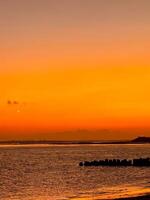 Orange sunset over Kelan Beach, Bali, with calm ocean waves and a scenic breakwater. A tranquil coastal scene with warm hues and serene atmosphere. photo