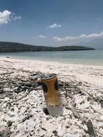 A close-up of a rebranded beverage can rests on the beach, its sleek design contrasts with rugged rocks, branches, and a vast blue ocean. Distant hills add natural calm, pure bliss. photo