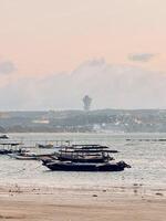 Traditional Fishing Boats at Kelan Beach Bali with Garuda Wisnu Kencana Statue in the Distance. Serene Coastal View at Sunset with Calm Ocean, Soft Clouds, and Distant Cityscape. photo