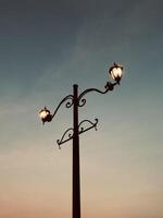 A vintage street lamp from a low angle warm light contrasts a sky shifting from dusk blue to soft orange. Twilight's nostalgic charm crowns this serene sentinel. photo