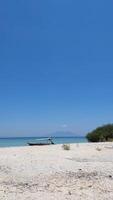 A serene view of Menjangan Beach, where one boat rests at shore. Crystal-clear blue waters mirror vivid bright sky and distant mountains, while soft white sand sets a calm scene. photo