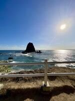 A breathtaking view of Papuma Beach at midday from an elevated vantage. The deep blue ocean meets a clear sky as a rugged rocky islet rises, framed by a wooden fence. A dream view. photo