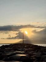 Serene Sunrise Over Rocky Jetty with a tall pole silhouetted with overcast cloud. Plengsengan Beach Banyuwangi, Bali Strait photo