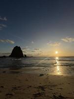 landscape view of Papuma Beach in Jember, featuring a rocky islet standing tall against the warm hues of the setting sun. The sky transitions from deep blue to orange. Papuma Beach Jember photo