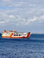 Large ferry sailing across the Bali Strait under a cloudy sky with deep blue ocean water. Passenger and vehicle transport ferry connecting Bali and Java, Indonesia, with open ramp and vibrant colors. photo