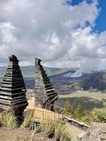 View of Mount Bromo with traditional Balinese split gate in the foreground under a blue sky with clouds. Stunning volcanic landscape, lush green hills, and volcanic smoke in East Java, Indonesia. photo