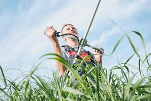 Man in overalls with weed trimmer mowing tall grass on sky background photo