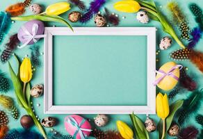 Easter Frame of yellow tulips, decorated eggs and feathers on turquoise background for greeting message photo