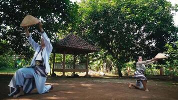 Two individuals performing a traditional dance in a rural setting, wearing traditional attire and conical hats. The background features greenery and a gazebo video