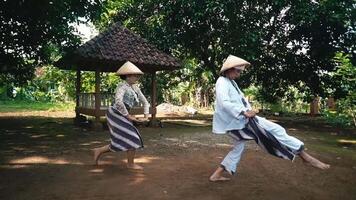 Two individuals performing a traditional dance in a rural setting, wearing traditional attire and straw hats, with a gazebo in the background surrounded by greenery video
