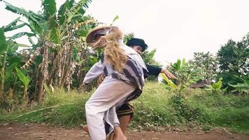 Two individuals engaged in a traditional martial arts practice in a rural setting, surrounded by greenery. One wears a black jacket and patterned sarong, while the other is in a white outfit video