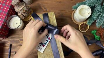 Woman tie black ribbon in bow on gift box in gold wrapper with tag. Label with embroidery in form of wreath, candles and other Christmas decor on wooden table. Top view of New Year's preparations. video