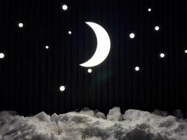 A luminous crescent moon and twinkling stars shine against a dark backdrop, casting a soft glow over the snow below. The minimalistic night sky installation creates a dreamy and serene winter photo