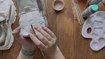 Top view of hands paint white boots with brush on wooden table. Palette with brown acrylic and other tools on desktop. Hobbies, creativity and handmade work at home. Designer's workshop. video