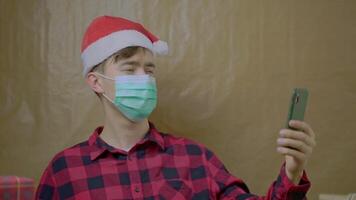 Man in medical mask waves at mobile phone camera. Guy in plaid shirt and Santa Claus hat is talking on chat with friends and relatives. Self-isolation during New Year holidays. video