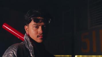 A young man with a serious expression holds a red baseball bat, wearing a shiny silver jacket and sunglasses. The background is dimly lit, creating a dramatic atmosphere video