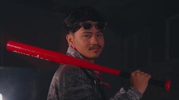 A young man holding a red baseball bat, dressed in a denim jacket and a red bandana. He has a serious expression and is positioned in a dimly lit environment, suggesting a dramatic or intense scene video