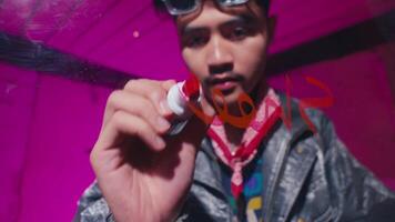 A young man wearing a leather jacket and goggles writes with a red marker on a glass surface. The background is a vibrant pink, creating a creative and artistic atmosphere video