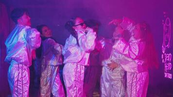 A group of young dancers performing energetically under colorful lights. One dancer in the foreground wears sunglasses and a shiny jacket, smiling broadly. The atmosphere is vibrant and lively video