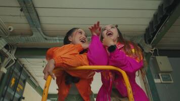 Two young women posing playfully in colorful jackets, one in orange and the other in pink, against an industrial backdrop. They are smiling and making fun expressions, showcasing a vibrant video