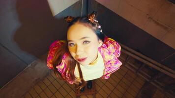 A young woman with colorful hair accessories and a vibrant jacket stands in a tiled room, viewed from above. The setting has an urban feel, with a focus on her unique style video