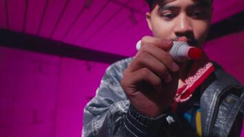 A young man with a mustache is focused on writing with a red marker. He wears a silver jacket and a colorful shirt, with a bandana around his neck. The background is illuminated in pink hues video