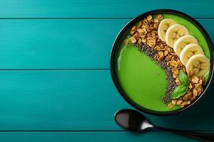 vibrant green smoothie bowl topped with banana slices, granola, and chia seeds, served in black bowl on teal wooden surface photo