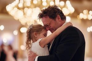 A father beams with pride as he dances with his young daughter at a luxurious evening event. Soft golden lights from chandeliers create a warm, enchanting atmosphere full of joy photo