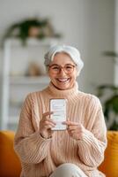 An elderly woman smiles while holding her smartphone, pointing at a digital app interface. The setting is bright and inviting, ideal for showcasing senior-friendly technology photo