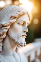 A close-up reveals a beautiful sculpted statue of Jesus. Soft sunlight filters through trees, creating a warm, divine glow that highlights his serene expression photo