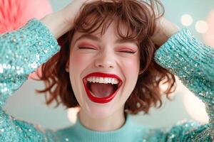 A fashionable woman with bold red lipstick, smiling broadly and laughing. Her hands are playfully placed on her head, eyes closed, against a blurred pastel colored backdrop photo