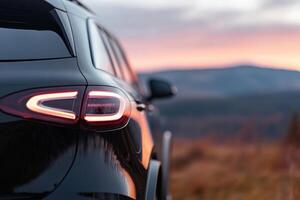 A black SUV is parked against a stunning backdrop of mountains as the sun sets, casting warm colors in the sky. The vehicle's taillight glows, enhancing the tranquil atmosphere photo