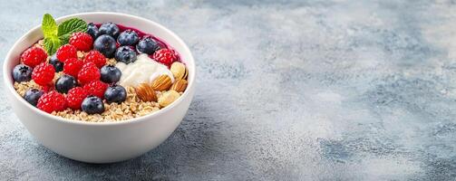 A vibrant bowl of yogurt topped with fresh raspberries, blueberries, and granola, garnished with a mint leaf on a textured blue background. photo