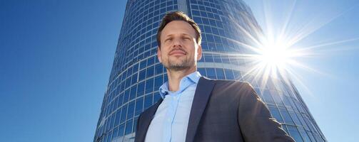 A confident man stands in front of a modern skyscraper, illuminated by sunlight, exuding professionalism and determination. photo