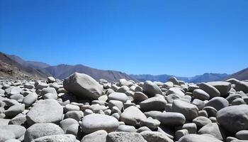 Smooth, rounded gray river rocks of various sizes scattered in the foreground of a mountainous landscape, with a clear blue sky above photo